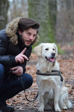 a man kneeling down next to a white dog in the woods with his tongue out