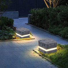 two stone benches sitting on top of a sidewalk next to plants and bushes at night