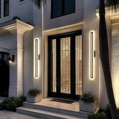 the front door of a modern home lit up at night with palm trees and potted plants