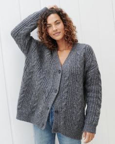 a woman wearing a gray cardigan sweater and jeans standing in front of a white wall