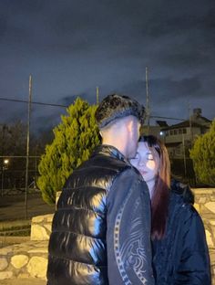 a man and woman standing next to each other in front of a stone wall at night