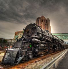 an old train sits on the tracks in front of a large building with a sky background