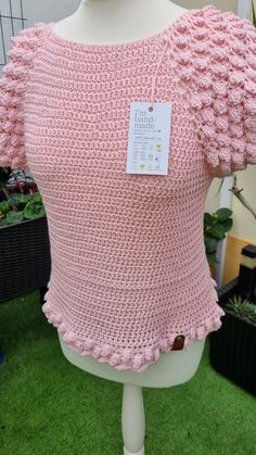 a crocheted pink top with ruffles on the shoulders is displayed in front of a white mannequin