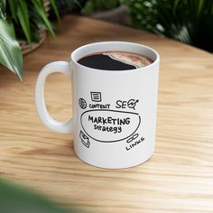 a white coffee mug sitting on top of a wooden table next to a green plant