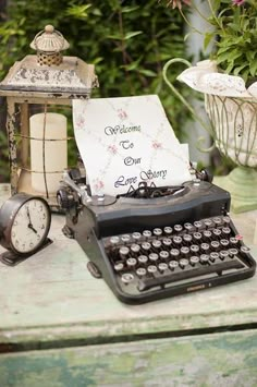 an old fashioned typewriter sitting on top of a table next to a clock and flowers