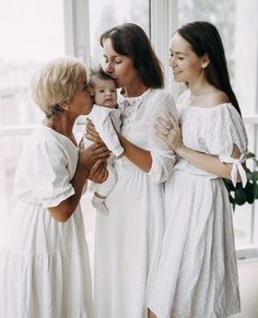 three women in white dresses are holding a baby and smiling at each other while standing next to a window