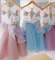 three dresses hanging on a rack in front of a window with sheer curtains and colorful tulle skirts