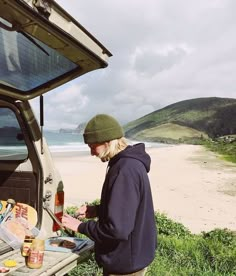 a man standing in front of an open car trunk with food on the beach behind him