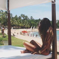 a woman sitting on top of a white bed next to the ocean and palm trees