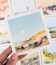 a hand holding up a small card with watercolor paintings on the wall behind it