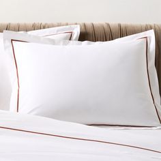 a bed with white sheets and brown piping on the pillowcase, along with two pillows