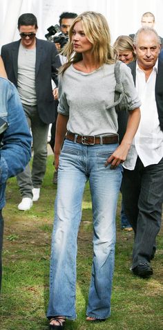 a woman in jeans is walking on the grass with other people behind her and another man standing next to her