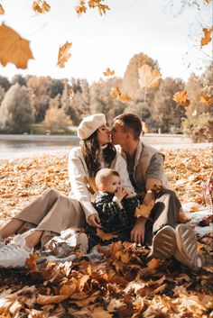 a man and woman kissing while holding a baby in the fall leaves near a lake