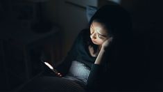 a woman sitting in the dark looking at her cell phone and reading something on her lap