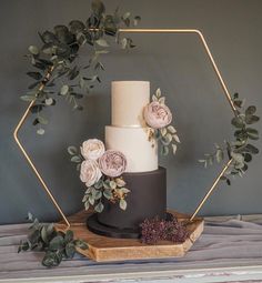 a three tiered cake with flowers and greenery on a wooden stand in front of a gray wall