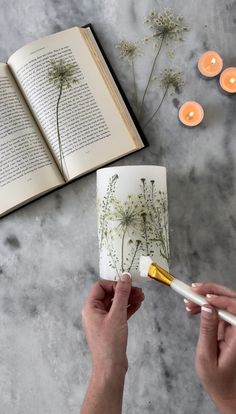 two hands holding an open book over a table with candles and flowers on it, while another hand holds a paintbrush