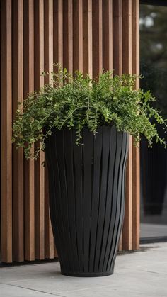 a large black planter with green plants in front of a wooden wall on the sidewalk