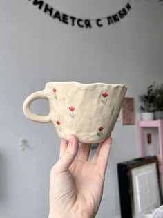 a hand holding a white cup with red flowers painted on it in front of a gray wall