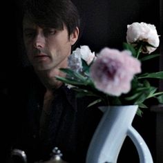 a man sitting at a table with a white vase and flowers in front of him