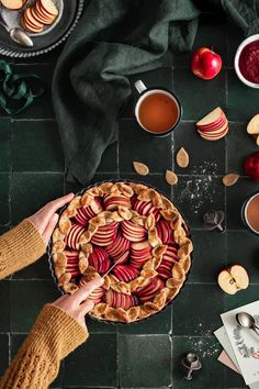 an apple pie on a table with apples and cups of tea next to the pie