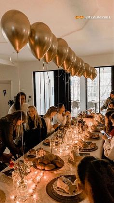 a group of people sitting around a long table with plates and candles on it, surrounded by balloons