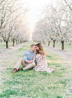 an engaged couple sitting on the grass in front of some trees with blossoming blossoms