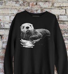 "This item is a Gildan 1800 personalized Unisex Sweatshirt portraying a cute sea otter design. It's a great present if you know someone that likes sea otters or you want to have it for yourself. It's clearly unique. You can have a lot of fun for parties or impress your friends with this gift.  Ideal for any situation, a unisex heavy blend crewneck sweatshirt is pure comfort. These garments are made from polyester and cotton. This combination helps designs come out looking fresh and beautiful. Th Graphic Print Long Sleeve Hoodie For Gift, Graphic Print Long Sleeve Hoodie As A Gift, Cute Sea Otter, Otter Design, Sea Otters, Sea Otter, Wild Animals, Print Pullover, Otters