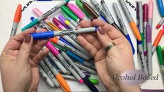 two hands are holding pens in front of many different colored pens on a white surface