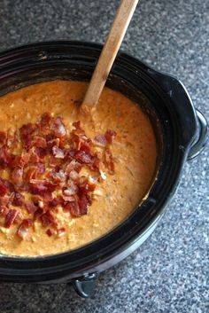 a black crock pot filled with soup and bacon on the top, next to a wooden spoon