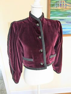 1976 Saint Laurent Rive Gauche Burgundy Velvet Bolero Jacket. This jacket closes with faceted black glass dome buttons, with an extra one sewn into the left side. The sleeves are leg of mutton style and it has a standing collar. The center front, collar, pockets, and sleeves are decorated with black trim. It is in excellent condition. Bust: 37" Waist: 37" Shoulders: 14.5" Sleeves: 30" Length: 17" Vintage Formal Blazer With Covered Buttons, Vintage Blazer With Covered Buttons For Formal Occasions, Vintage Outerwear With Stand Collar And Buttons, Vintage Outerwear With Historical Design For Costume, Vintage Costume Outerwear With Historical Design, Vintage Blazer With Covered Buttons, Vintage Evening Outerwear With Buttons, Vintage Formal Blazer With Snap Buttons, Vintage Blazer With Snap Buttons For Formal Occasions