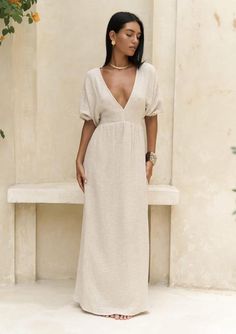 a woman standing in front of a bench wearing a white dress and gold necklaces