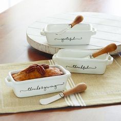 three white dishes with bread in them sitting on a table next to two serving utensils