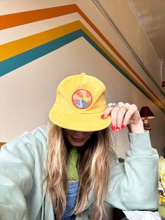 a woman with long hair wearing a yellow hat and blue jacket sitting in front of a colorful wall