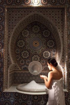 a woman in a white dress is looking at something on a shelf with an intricately designed wall behind her