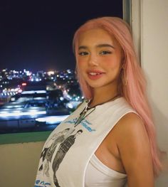 a woman with pink hair standing next to a window in front of a cityscape