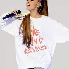 a woman with long hair holding a microphone and wearing a white shirt that says one love manchester