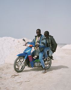 two men are sitting on a motorcycle in the desert, one is wearing a blue jacket