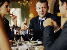 three people sitting at a table with wine glasses in front of them and one person pointing to the side