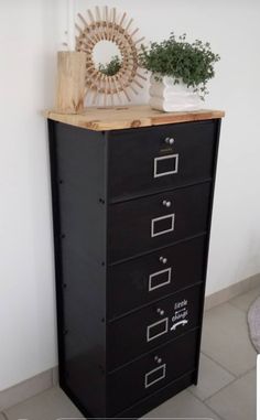 a black filing cabinet with five drawers in front of a white wall and potted plant on top