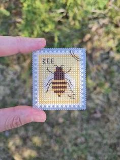 a person holding up a small needle - embroidered square with a bee on it's side