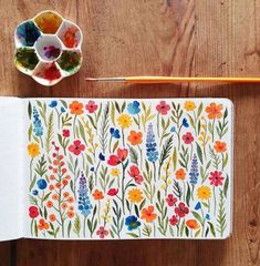 an open notebook with flowers painted on it next to a paintbrush and watercolor pencils