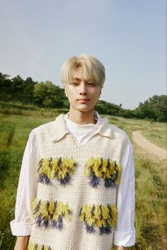 a young man standing in the grass wearing a white sweater with yellow and blue flowers on it