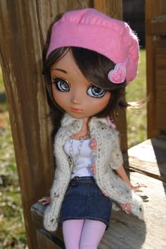 a doll sitting on top of a wooden bench wearing a pink hat and sweater with flowers