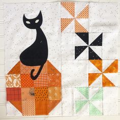 a black cat sitting on top of a patchwork piece of quilted material with orange and green squares