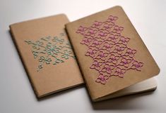 two notebooks with embroidered designs on them sitting next to each other, one opened and the other closed