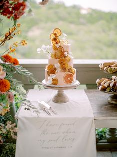 a three tiered wedding cake with oranges on top and flowers in the background