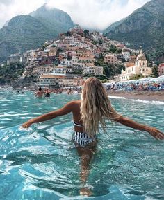 a woman standing in the ocean with her arms spread out and looking at the beach