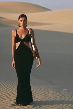 a woman standing in the sand wearing a black dress with cutouts on it's sides