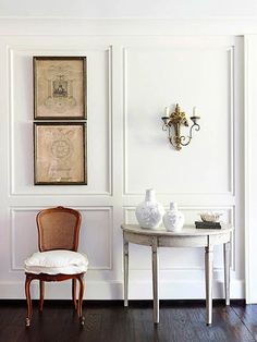 a chair and table in a room with white paneling on the walls, two framed pictures above them