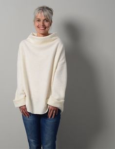 a woman standing in front of a gray wall wearing a white sweater and jeans with her hands on her hips